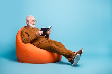 portrait of his he nice attractive focused cheerful cheery wise smart clever bearded grey-haired man sitting in bag chair reading academic book isolated over blue pastel color background