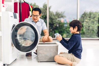 family asian father and kid little boy son having fun doing household chores doing laundry dirty clothes into the washing machine together in laundry room at home
