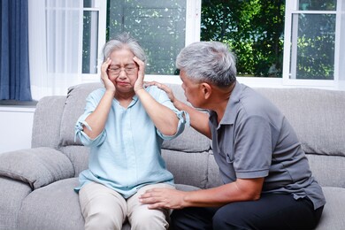 an elderly asian couple sitting on the sofa at home a husband takes care of a wife who has a headache and is sick. senior health concept