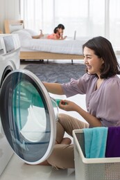 smiling asian woman sitting on the floor loading clothes into a laundry machine for her routine housework on holiday at home with soft background of a young girl lying on a bed.