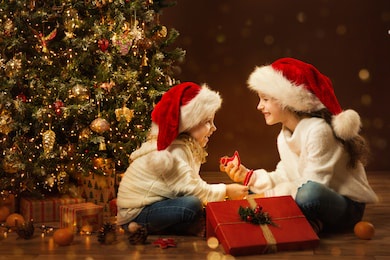 christmas child girl opening box and giving gift toy to sister. happy children in santa hat next to xmas tree. fairy magic interior, indoor lights
