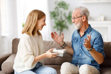excited young daughter smiling and giving gift box to cheerful aged man while sitting on couch during holiday celebration
