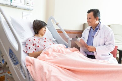 confident asian medical doctor explaning to cheerful female patient on medical condition lying on bed in hospital ward