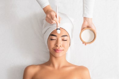 beauty treatments. cosmetologist applying clay mask on asian woman face at spa salon, relaxed korean lady with towel on head enjoying wellness day at beauty center, top view with free space