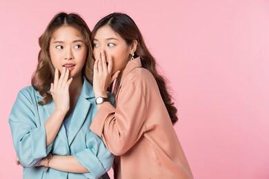 two pretty women whispering in ear a secret story. portrait isolated on pink studio background. 