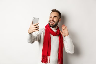 handsome bearded man video calling friends, saying hello and waving hand at mobile phone, standing in sweater and red scarf against white background