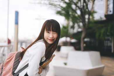 college people lifestyle on day time concept. portrait of young adult asian student woman. hold notebook and wear backpack at outdoor. city and river blur background.