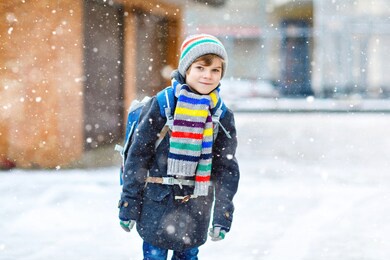 little school kid boy of elementary class walking to school during snowfall. happy child having fun and playing with first snow. student with backpack or satchel in colorful winter clothes.