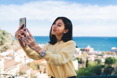 cheerful asian female in casual clothes and wristwatch standing on balcony and taking selfie on smartphone against seascape during sunny day
