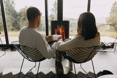 stylish couple in sweaters with warm cups of tea relaxing at modern black fireplace with view on mountains. cozy warm family moments at cold season. young family on vacation