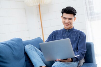 young asian businessman smile and work from home with laptop computer online to internet on sofa in living room, freelance man using notebook on couch with comfort, new normal, lifestyle concept.