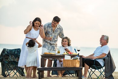 group of happy multi generation asian family enjoy dinner party together on the beach at sunset. parents with senior grandparents and cute child girl relax and having fun on summer holiday vacation