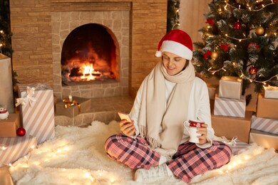 photo of young lady sitting on floor and holding cup with tea or coffee, looking at mobile phone's screen, wears white pullover and checkered pants, in decorated x-mas living room.