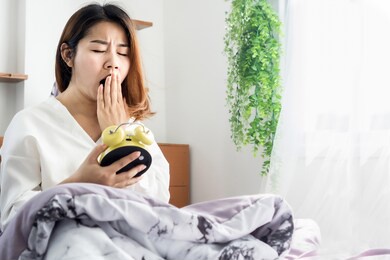 asian woman yawning bed hand holding alarm clock feeling tired and sleepy 