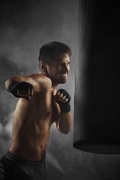 aggressive boxer in black boxing wraps punching in boxing bag on dark background with smoke