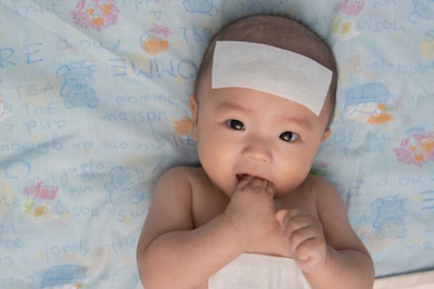 unhappy asian chinese little baby child sick suffering heat from fever with cool fever jel pad on forehead and body