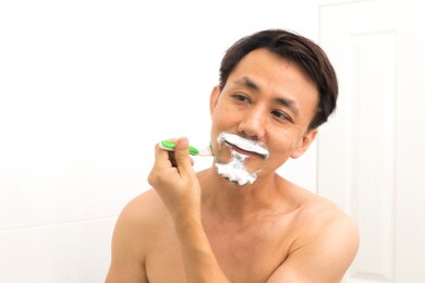 middle aged asian man putting on shaving cream and shave his beard in the bathroom.