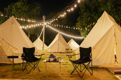 group outdoor camping teepee tent and night light with two empty chairs with picnic table and accessories in the forest. glamping camping tent in the forest under night sky.