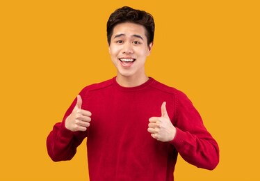 everything is fine. portrait of happy asian man showing thumbs up, doing approval gesture with fingers and both hands, smiling, showing that he is good, isolated over orange studio background