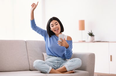 chill and lifestyle concept. exited asian woman wearing wireless earphones listening to favorite song, holding mobile phone, sitting on sofa and dancing in living room, raising hand and finger up