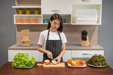 young asian woman hands cooking and cutting an apple on wooden cutting board preparing healthy food, holding knife and slicing fruits on table in kitchen