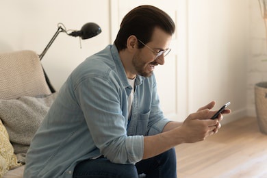 close up smiling man wearing glasses using smartphone, chatting online with friends, enjoying leisure time with gadget, weekend, playing mobile device game, having fun, sitting on cozy couch at home