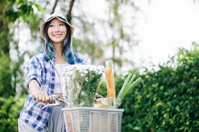 smiling lovely young chinese woman riding on bicycle with flower bouquet and food in front basket