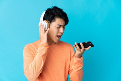 young chinese man isolated on blue background listening music with a mobile and singing