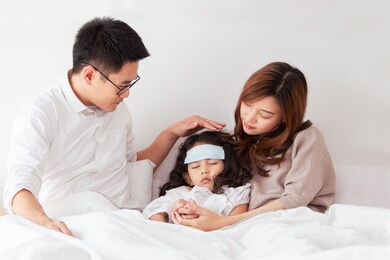 asian sick girl sleeping on the bed with fever gel pack on her forehead. father and mother take care of their little young daughter who got a fever from flu virus. sick little kid laying on the bed.
