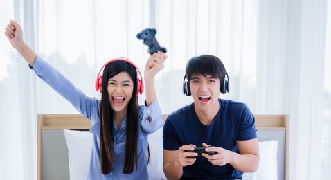 happy young couple wear headphone and holding joystick playing video game on console to fun. asian couple wearing earphone and sitting with relaxation by technology play station on bed in bedroom.