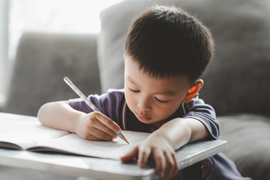 asian boy doing homework with the intention. child boy holding pencil writing, a boy drawing on white paper at the table, elementary school and home schooling, distance education concept.