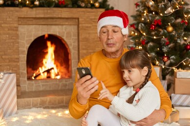granddaughter sitting with her grandfather on floor on soft carpet near fireplace and decorated fir tree, granddad holding smart phone in hands, family looking at device screen.