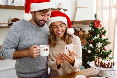 charming cheerful couple in christmas hats smiling and using cellphone in cozy kitchen