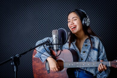 happy cheerful pretty smiling of portrait a young asian woman vocalist wearing headphones with a guitar recording a song front of microphone in a professional studio