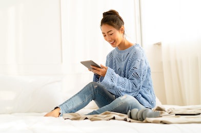 beautiful young asian woman wearing knitted sweater sitting in comfort using tablet at cozy home atmosphere on the white bed. lifestyle, comfort and technology concept. fashion, autumn, winter.