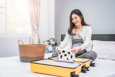 beautiful asian pregnant woman holding a tiny cute baby clothe and looking at it joyfully, siting on bed folding and putting baby clothing in yellow suitcase. resting smiling and relaxing in bedroom