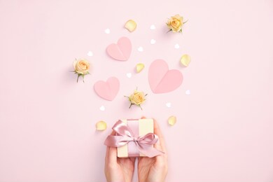 valentines day gift box in female hands, paper hearts, flowers and decorations on pink background. flat lay, top view.