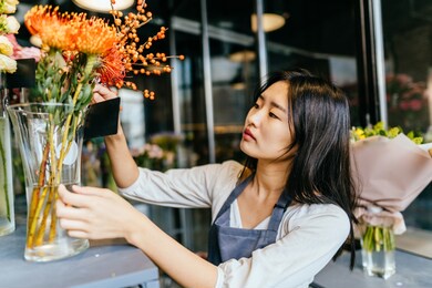 portrait of a young smiling asian florist in grey apron standing at the counter focusing on creating a bouquet for a client, flower shop interior. floristics, business, decoration concept.