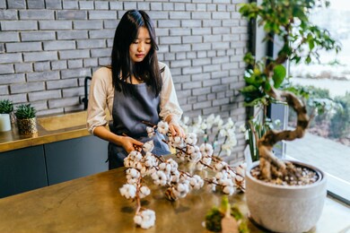 asian female florist or saleswoman wearing grey apron with raw cotton flowers in modern loft flower shop interior. creative work at flower shop. floristics business, winter decoration concept.