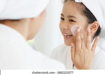 mother adding treatment cream on the cheek to young and cute asian girl with spa dress and head covered with a white towel.