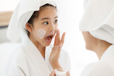 mother adding treatment cream on the cheek to young and cute asian girl with spa dress and head covered with a white towel. the kid's face feels excited and joyful.
