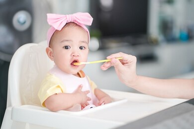 asian cute baby girl in a pink bib and hair bow sitting on a white high chair. her mother giving healthy food by spoon in the morning at home