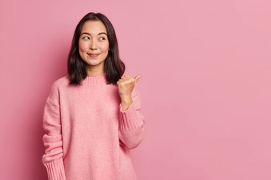 charming pleased young asian woman with rouge cheeks points aside on copy space shows advertisement smiles gently wears casual jumper isolated over rosy background. attention to this promotion