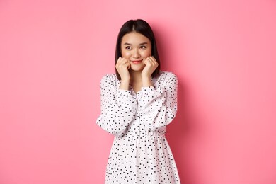 cute and shy asian girl blushing, touching cheeks and looking left at copy space silly, standing against pink background