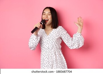 happy asian woman dancing and singing in microphone, performing at karaoke, standing over pink background