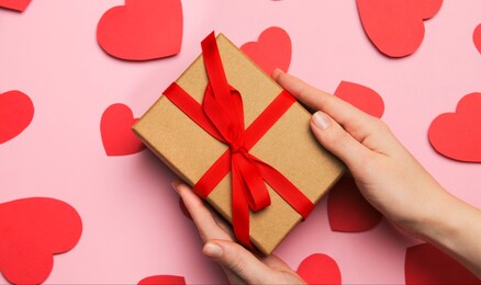 hands hold gift box present with red bow for valentine's day, declaration of love. composition with red hearts for valentine's day on a pale pink background. flat lay. love and relationships.