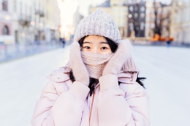 outdoor portrait of interested asian woman having fun in holidays. korean girl wearing hat and gloves covered her face with knitted scarf in cold sunny winter day.