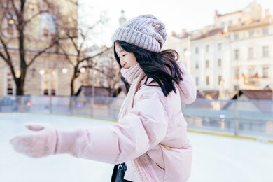 happy asian woman going to ice skating outdoor. korean female dressed in powder pink jacket and warm knitted hat.healthy lifestyle and sport concept at city square. winter weather at sunny day.