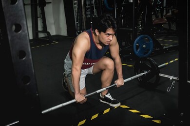 a handsome young asian male kneels on one leg, grasping the barbell and with an intense, motivated look.