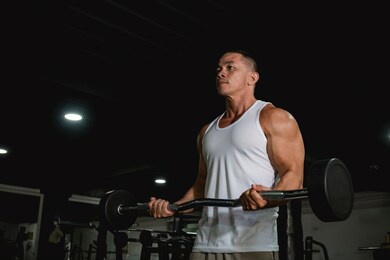 a well built and fit asian man in a white tank top does some barbell curls at the gym. an active 40 year old with healthy lifestyle.